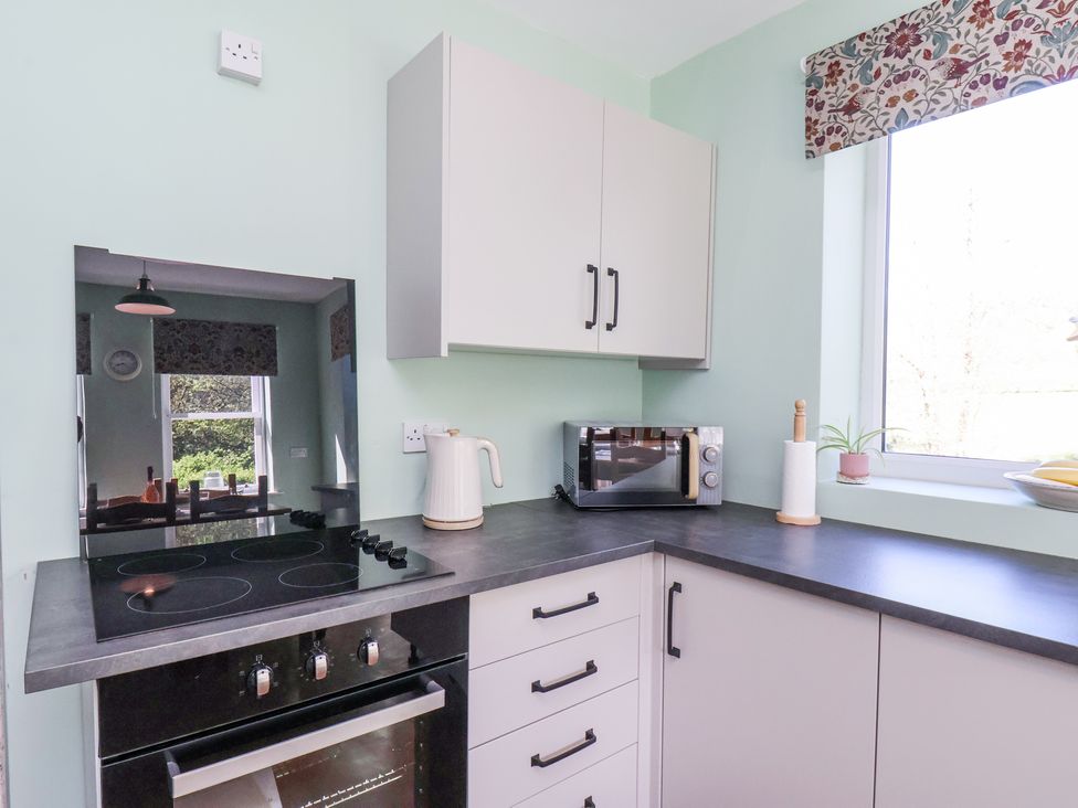 A kitchen corner with a black electric stove and oven white cabinets a microwave kettle paper towel holder and a window with floral blind at Whinstone Cottage Esk Valley near Grosmont