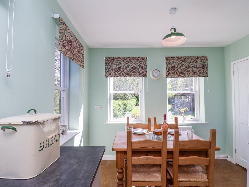 A dining area with a wooden table and chairs by three windows with floral blinds and a bread box on the counter at Whinstone Cottage in Esk Valley near Grosmont