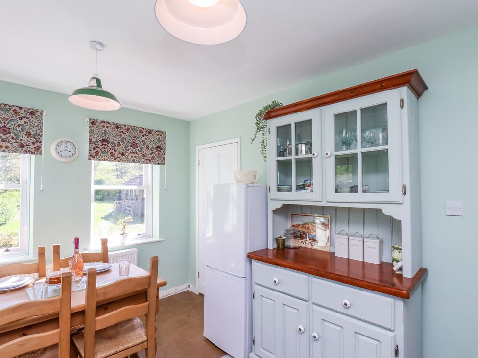A kitchen with a wooden dining table set by two windows and a white cabinet with glass doors at Whinstone Cottage Esk Valley near Grosmont