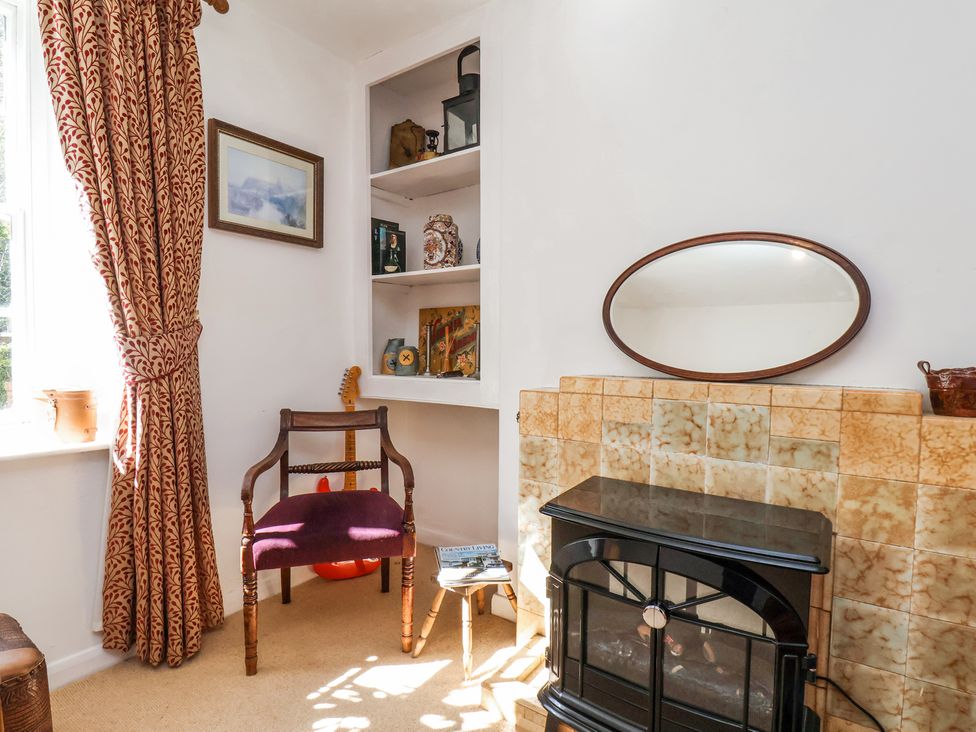 A corner of a room with a wooden chair, a small table with magazines, a guitar, shelves with decorative items, an oval mirror, and a tiled fireplace at Whinstone Cottage in Esk Valley near Grosmont