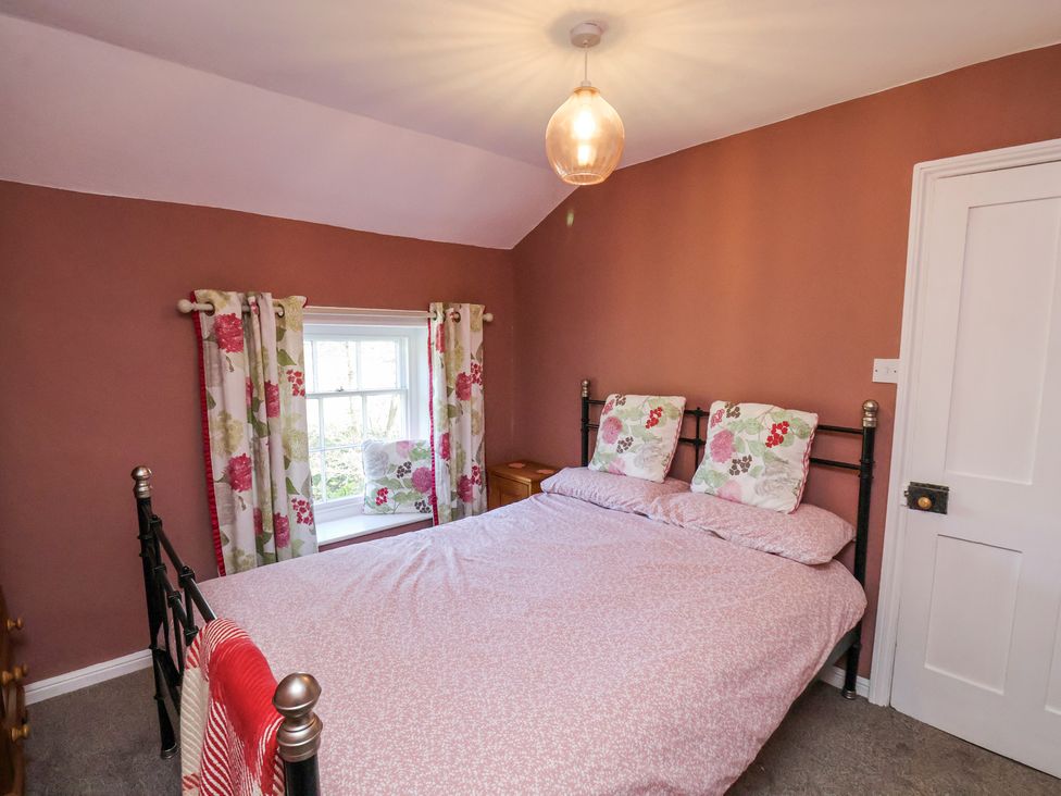 A bedroom with a double bed with floral pillows and bedding next to a window with floral curtains at Whinstone Cottage in Esk Valley near Grosmont