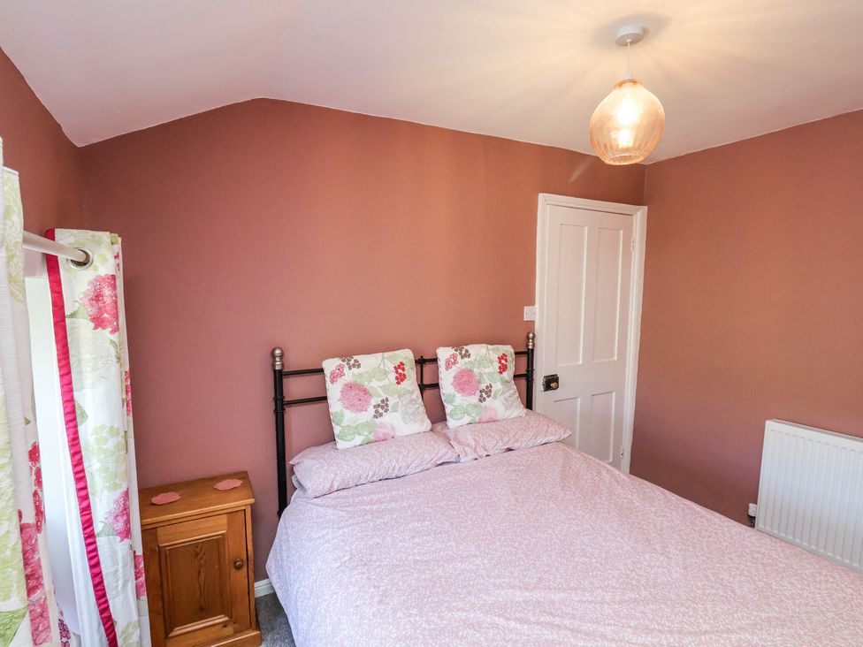 A bedroom with a double bed with floral pillows a bedside cabinet a door and floral curtains at Whinstone Cottage Esk Valley near Grosmont