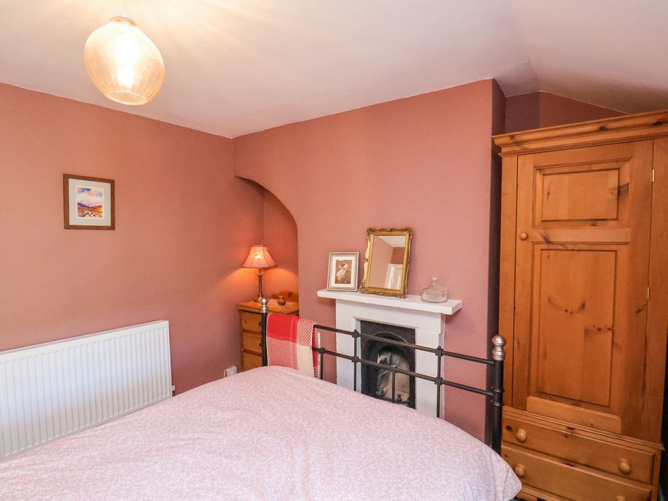 A bedroom with a bed a wooden wardrobe a chest of drawers a lamp and a small fireplace at Whinstone Cottage Esk Valley near Grosmont
