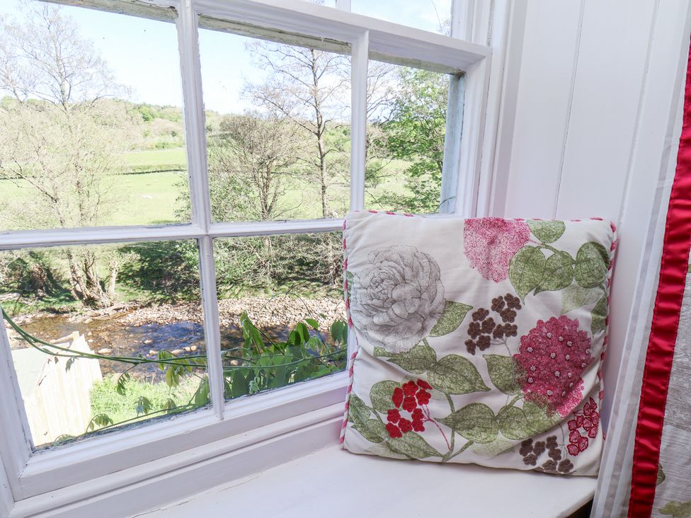 A cushion with floral pattern on a white window sill overlooking a river and trees at Whinstone Cottage in Esk Valley near Grosmont