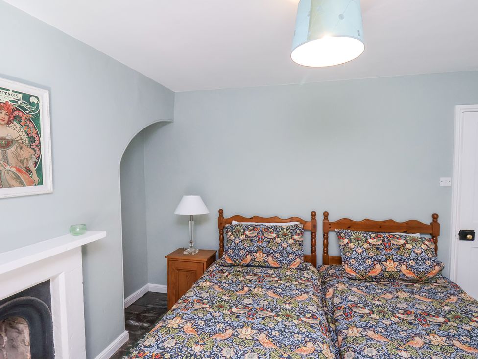 A bedroom with two single beds with patterned bedding a wooden nightstand with a lamp a fireplace and a framed picture on the wall at Whinstone Cottage Esk Valley near Grosmont