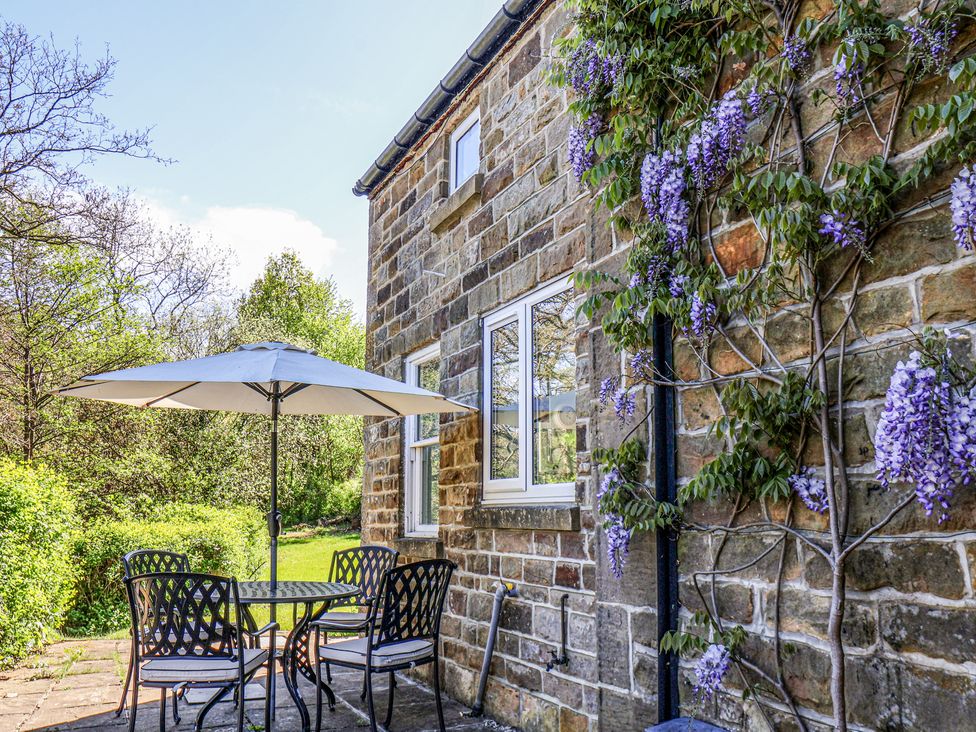 An outdoor patio with a table four chairs and a large umbrella next to a stone wall with climbing purple flowers at Whinstone Cottage in Esk Valley near Grosmont