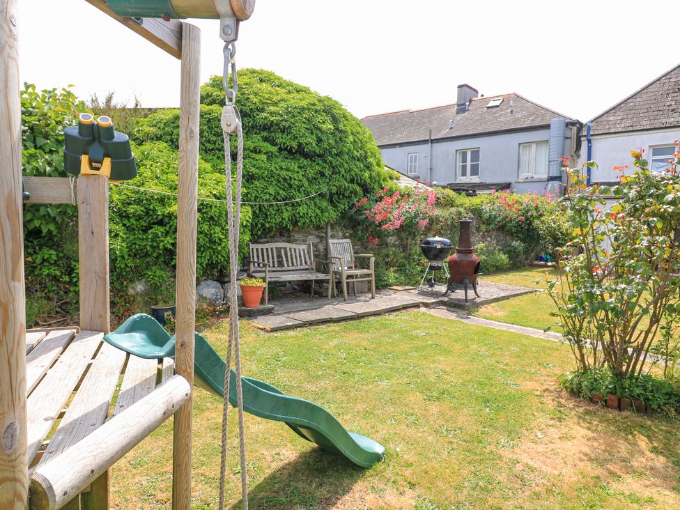 A backyard with a wooden play structure with a slide and binoculars a grassy lawn wooden benches a barbecue grill and a chiminea surrounded by bushes and flowers at Joymead in Malborough