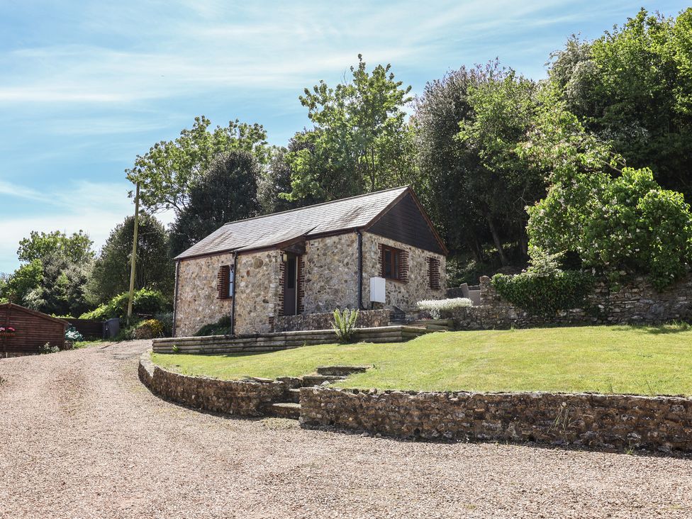 A stone cottage with a gravel driveway and grass lawn surrounded by trees at Megs Roost in Shute near Colyton