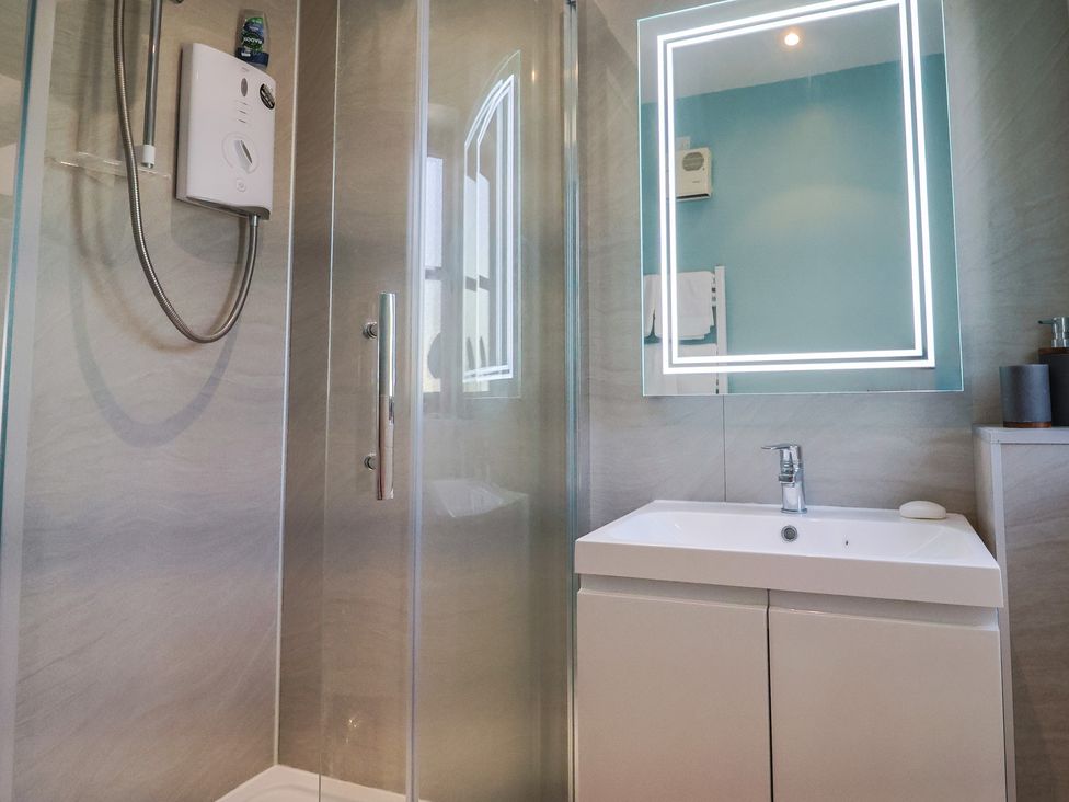 A bathroom with a shower enclosure, a wall-mounted electric shower, a sink with a cabinet, and an illuminated mirror at Megs Roost Shute near Colyton