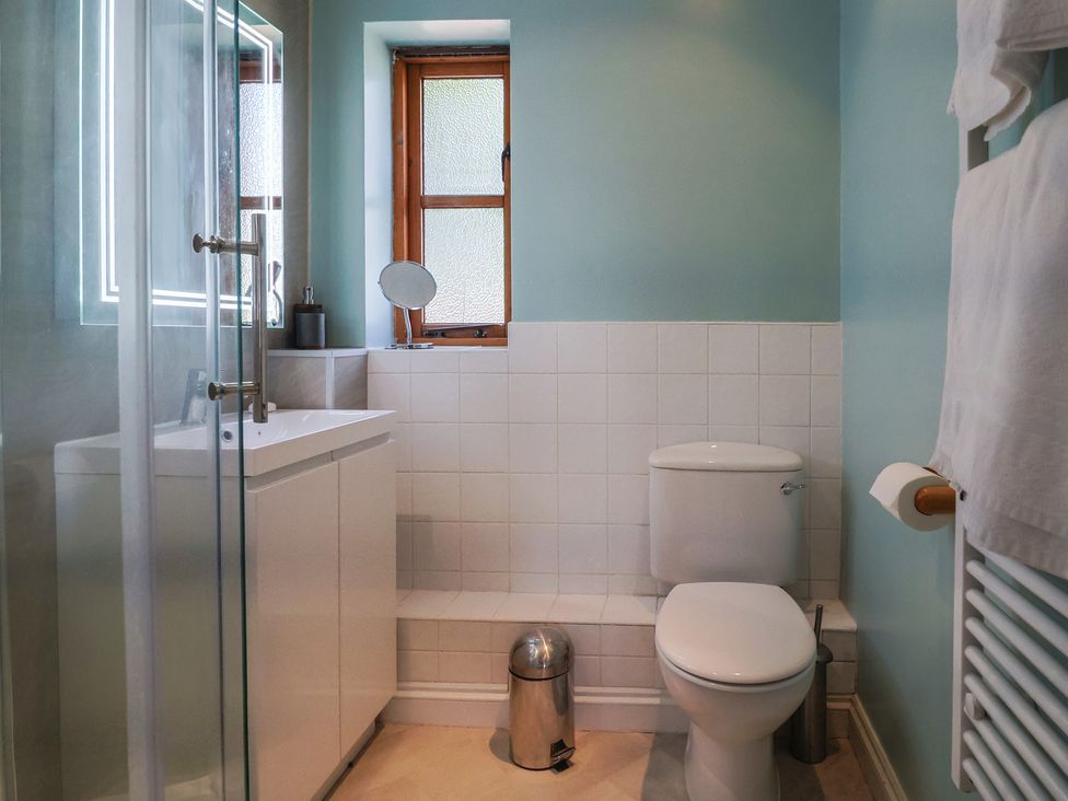 A bathroom with a toilet, sink, small mirror, shower, trash bin, and towel rack at Megs Roost in Shute near Colyton