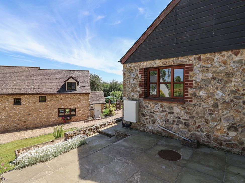 An outdoor stone patio area next to a stone and brick building with a window and a neighboring stone building in the background at Megs Roost in Shute near Colyton