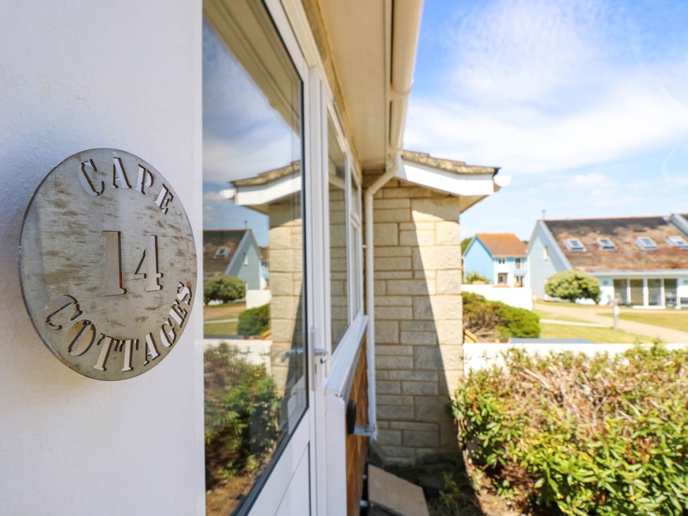 A white wall with a round metal sign reading cape 14 cottages next to a glass door with houses and shrubs in the background at Cape 14 in Yarmouth