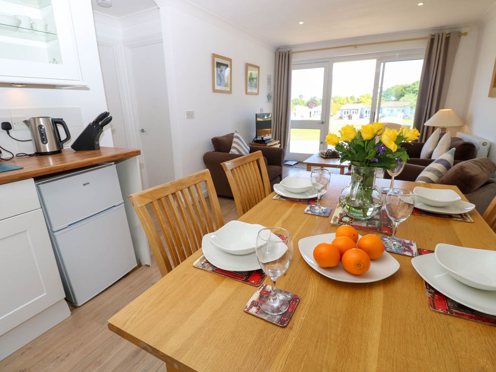 A dining table set with plates glasses bowl of oranges and a vase of yellow roses next to a kitchen with a kettle knife block and fridge and a living room with sofas and a TV at Cape 14 in Yarmouth