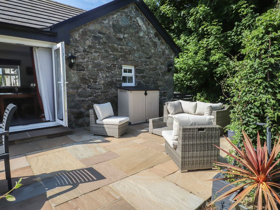 An outdoor patio with cushioned rattan chairs a stone wall and open doors at Annexe at Ty Moel Isaf in Pengorffwysfa near Amlwch