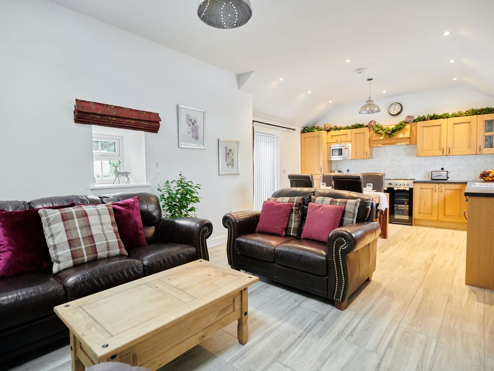 A living area with brown leather sofas red and plaid cushions wooden coffee table and an open kitchen with wooden cabinets at Annexe at Ty Moel Isaf in Pengorffwysfa near Amlwch