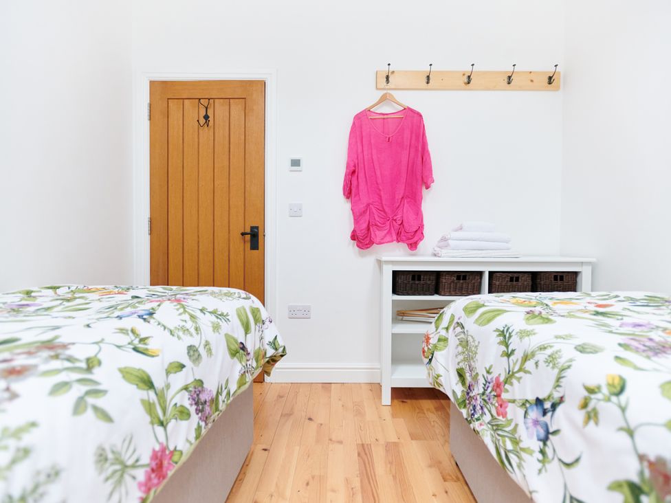 A bedroom with two beds with floral bedding a wooden door a white storage unit with baskets and folded towels and a pink shirt hanging on wall hooks at Annexe at Ty Moel Isaf in Pengorffwysfa near Amlwch