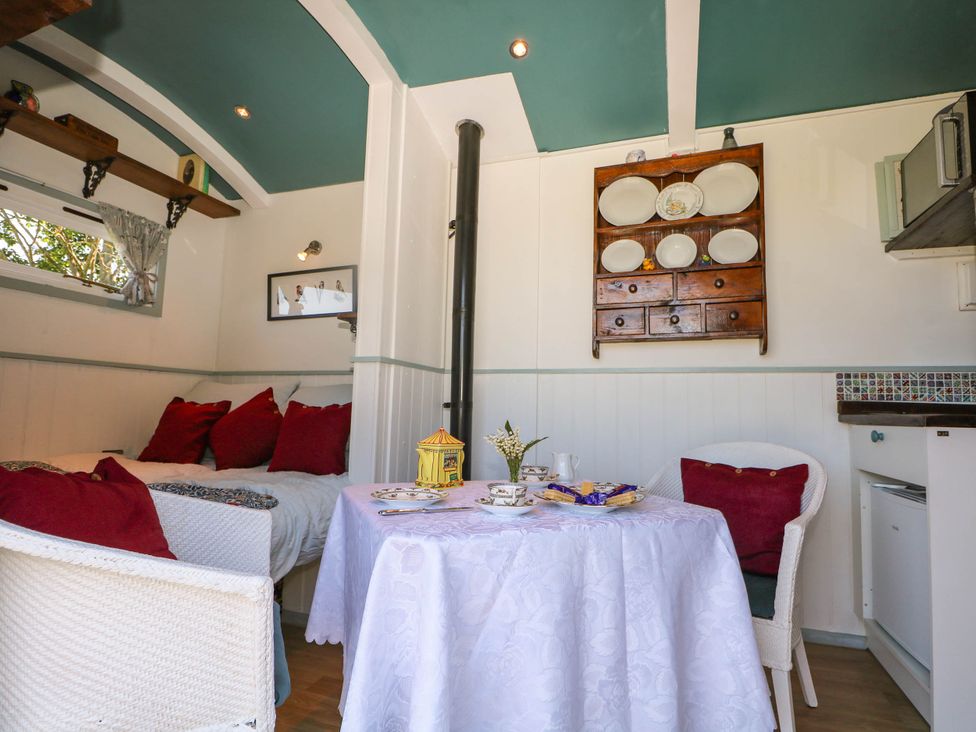 A dining area with a small table covered with a white tablecloth two white chairs with red cushions a bench with red cushions a wooden shelf with plates and drawers and a window with a curtain at Songbirds in St Michaels Kent