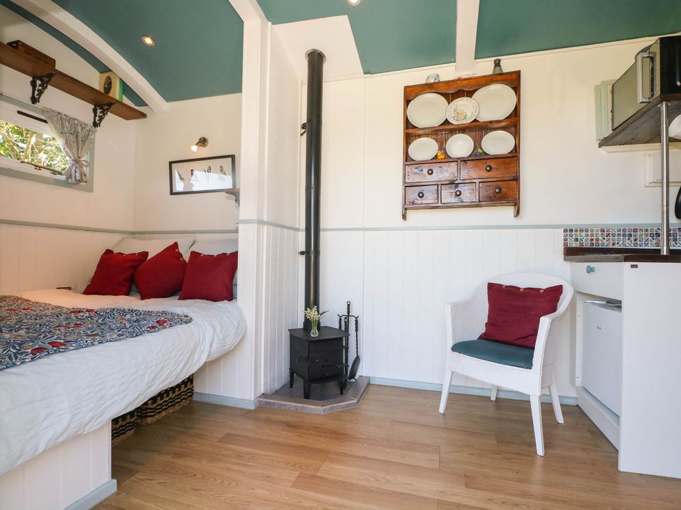 A small room with a bed with red pillows a white armchair with cushions a wood stove and a wooden shelf with plates on the wall at Songbirds in St Michaels Kent
