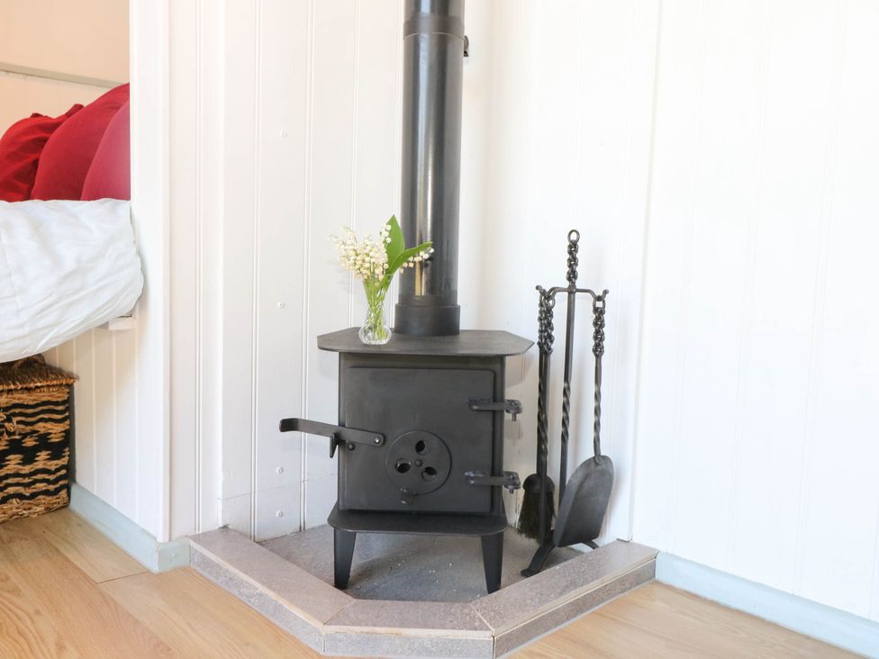 A black metal wood burning stove with a chimney pipe a small vase with flowers on top and fireplace tools next to it in a corner with white paneled walls and wood flooring at Songbirds in St Michaels Kent