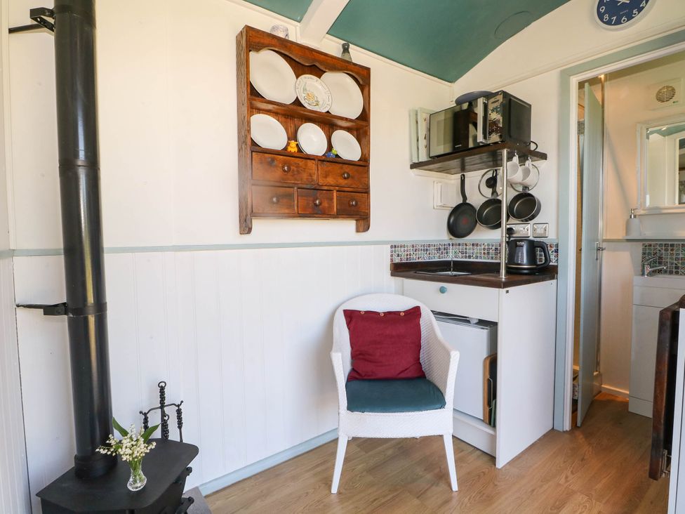A small kitchen area with a white chair with red and blue cushions a stove a wall-mounted wooden cupboard with plates and a microwave on a shelf at Songbirds in St Michaels Kent