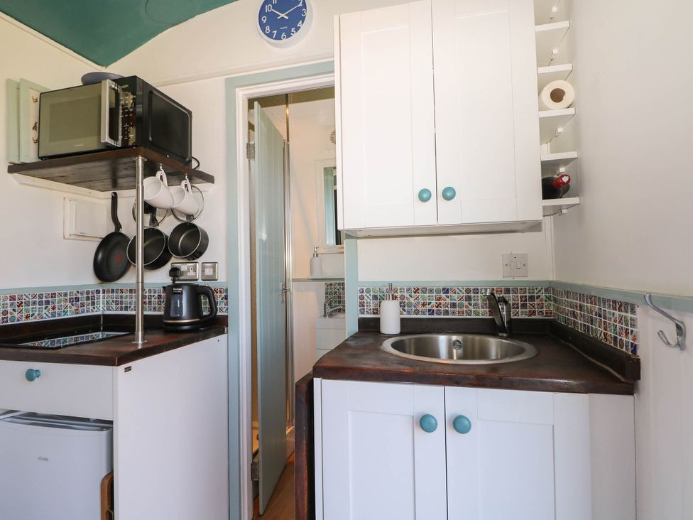 A small kitchen with a sink, microwave, kettle, hanging pans, and a door leading to a bathroom at Songbirds in St Michaels Kent