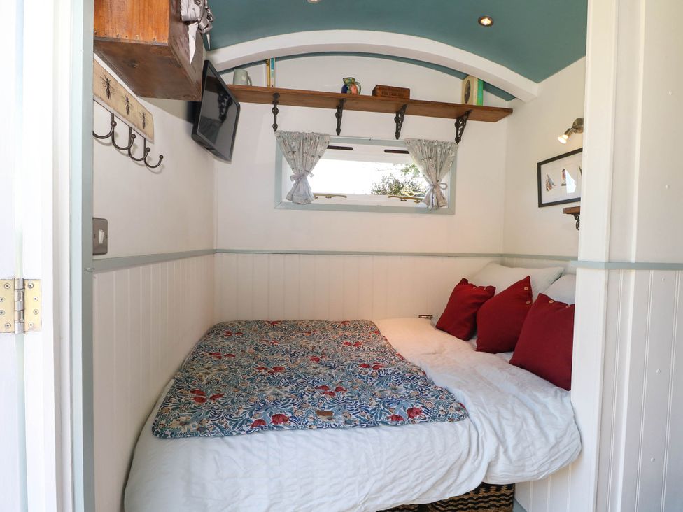 A small bedroom with a bed covered in red pillows and patterned bedding under a window with curtains at Songbirds in St Michaels Kent
