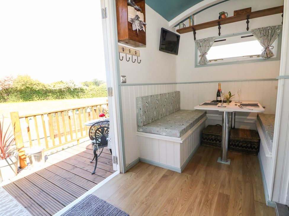A small dining area with built-in bench seats around a table set with a bottle and glasses inside a room with an open door leading to a wooden deck with a small black metal table and chairs at Songbirds in St Michaels Kent