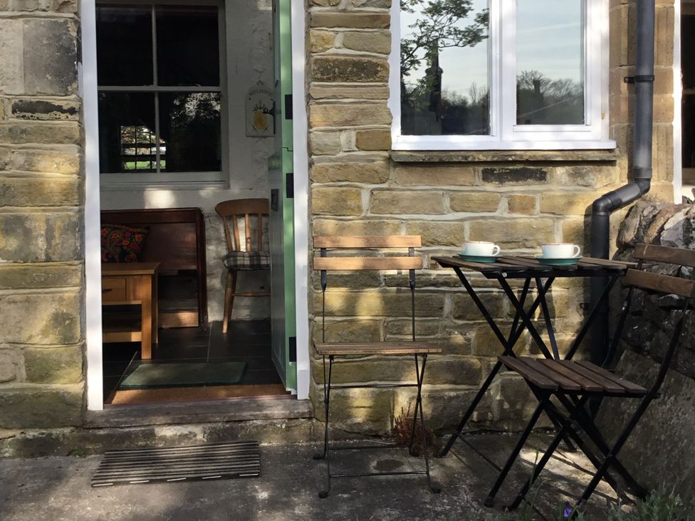 A stone exterior wall with an open door leading inside a room with a wooden chair and table and an outdoor seating area with a small table and two chairs with cups on the table at Doffers Cottage in Airton