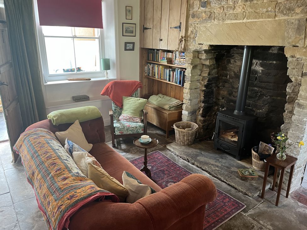 A sitting room with a sofa and armchair near a stone fireplace with a wood stove at Doffers Cottage in Airton