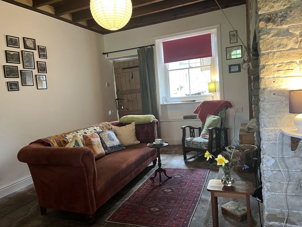 A room with a brown sofa, an armchair, a side table with a vase of flowers, a window with red blinds and a wooden door at Doffers Cottage in Airton