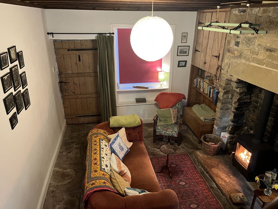 A living room with a brown sofa and patterned cushions a stone fireplace with a wood stove a wooden door and book shelves at Doffers Cottage in Airton