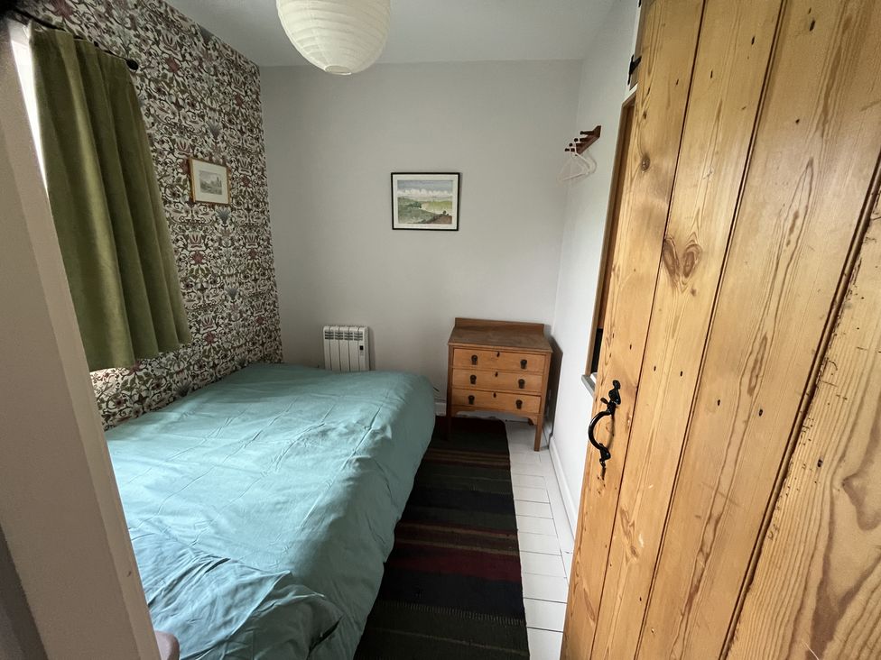 A small bedroom with a bed covered by a green duvet a wooden chest of drawers a patterned wallpapered wall an olive green curtain and a wooden door at Doffers Cottage in Airton