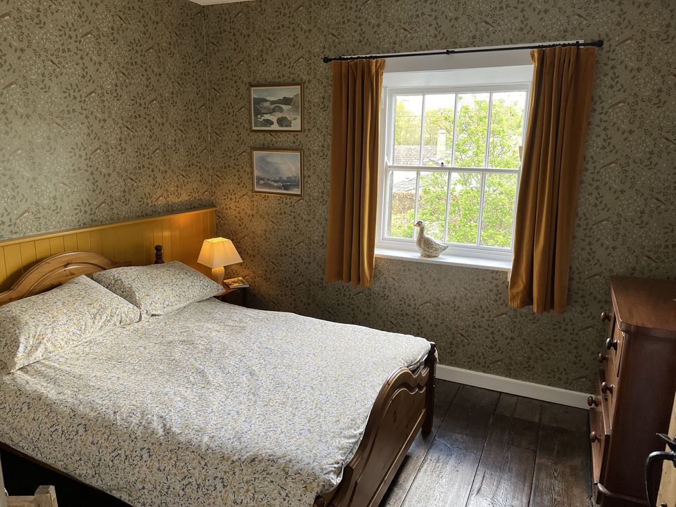 A bedroom with a wooden bed, patterned bedspread, a window with mustard curtains, a lamp on a bedside table, two framed pictures on the wall, and a wooden chest of drawers at Doffers Cottage in Airton