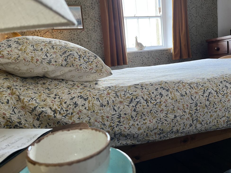 A bedroom with a bed covered in floral bedding a cup and book on a table next to the bed and a window with curtains at Doffers Cottage in Airton