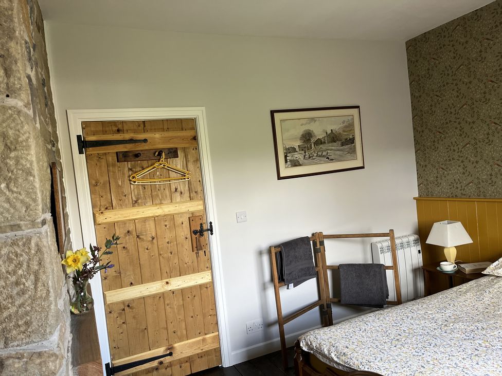 A bedroom corner with a wooden door a towel rack a bed a lamp on a bedside table and a framed painting at Doffers Cottage in Airton