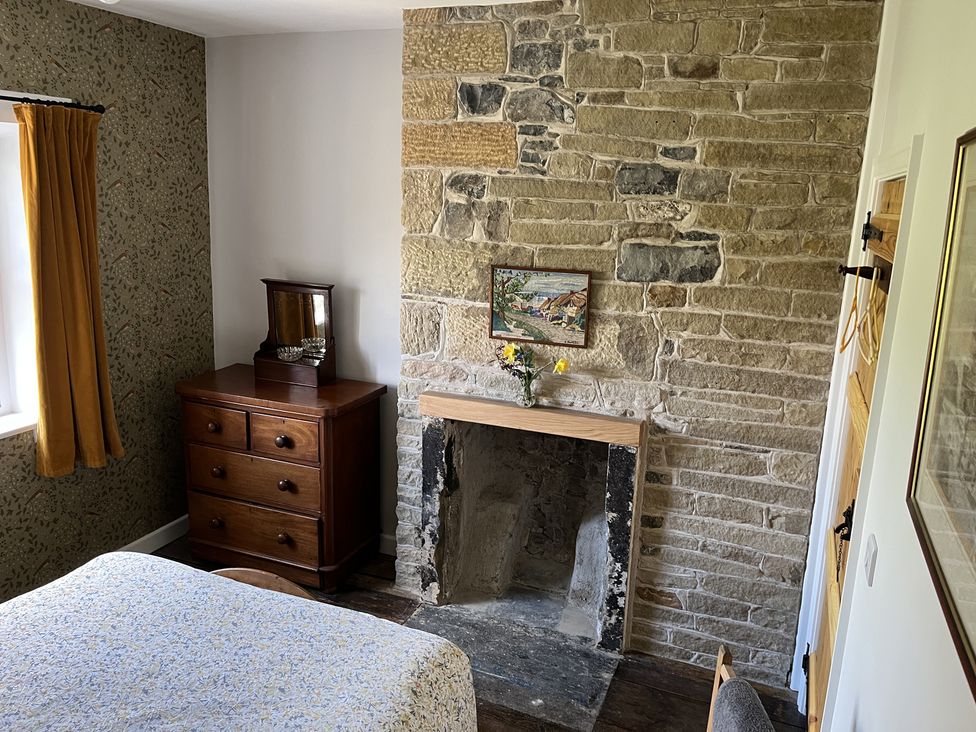 A bedroom with a stone fireplace wooden dresser with a mirror and a bed with patterned bedspread at Doffers Cottage in Airton