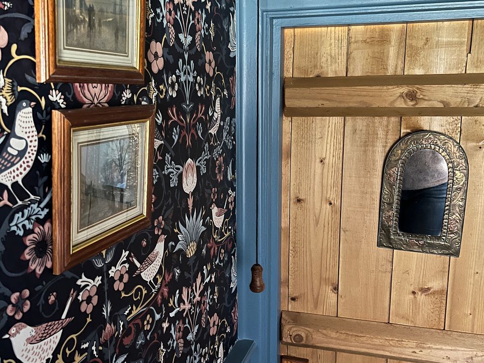 A decorative wall with framed pictures and patterned wallpaper next to a wooden door with a small ornate mirror at Doffers Cottage in Airton