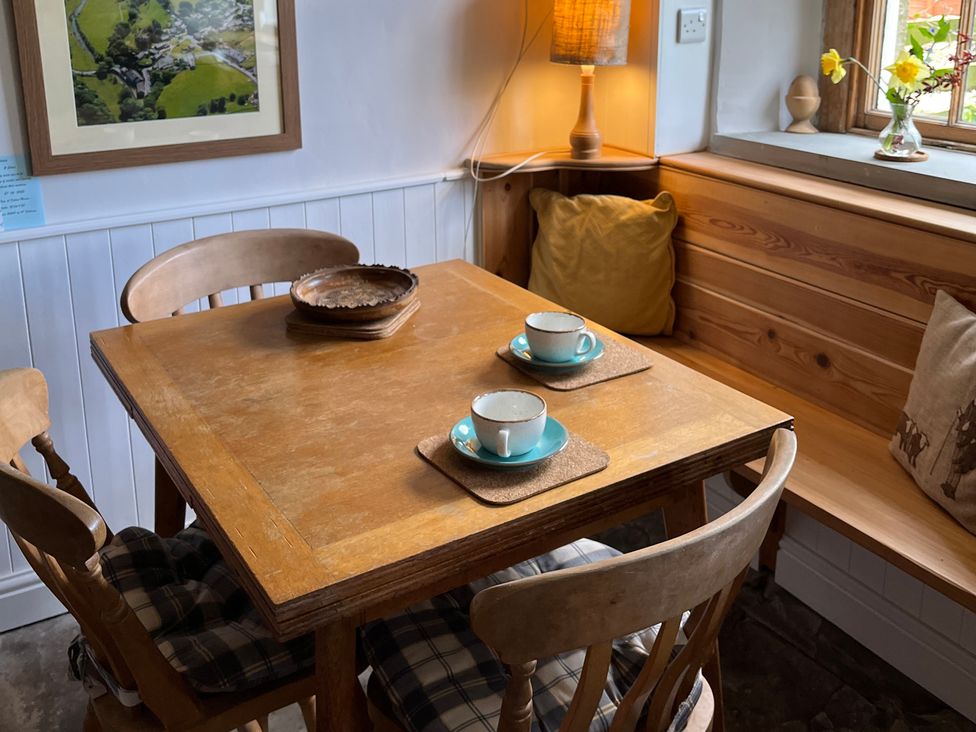 A wooden dining table with four chairs and a built-in bench with cushions in a corner next to a window with flowers at Doffers Cottage in Airton