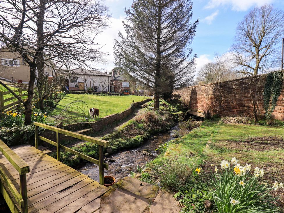 A garden with a bridge over a stream and a dog at Yew Tree Cottage in Mealsgate near Ireby