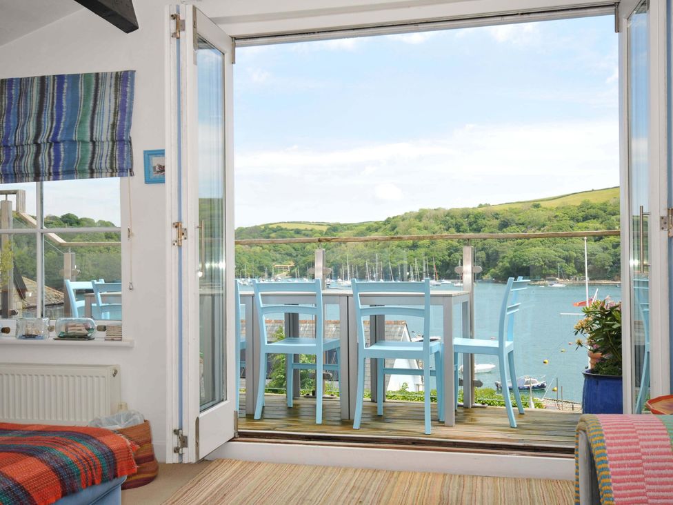 A room with an open glass door leading to a balcony with blue chairs and a table overlooking a river and green hills at 5 Esplanade in Fowey