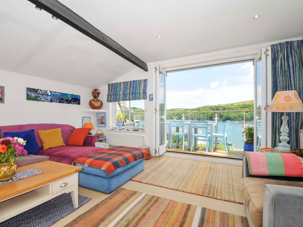 A living room with a purple sofa, colorful cushions, striped rugs, a coffee table, and an open door leading to a balcony with blue chairs overlooking water at 5 Esplanade in Fowey