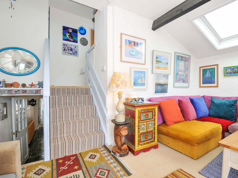 A living room with a yellow sectional sofa with colorful cushions a wooden chest with a table lamp and framed pictures on the walls at 5 Esplanade in Fowey