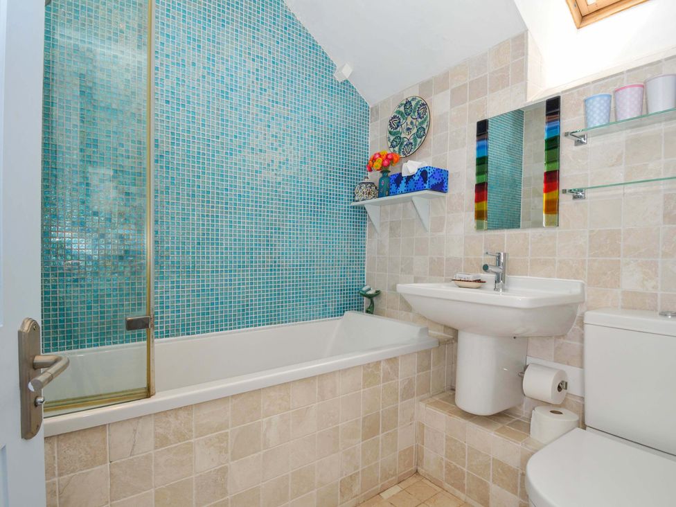 A bathroom with a bathtub tiled in blue and beige a sink a toilet shelves with cups and a mirror at 5 Esplanade in Fowey