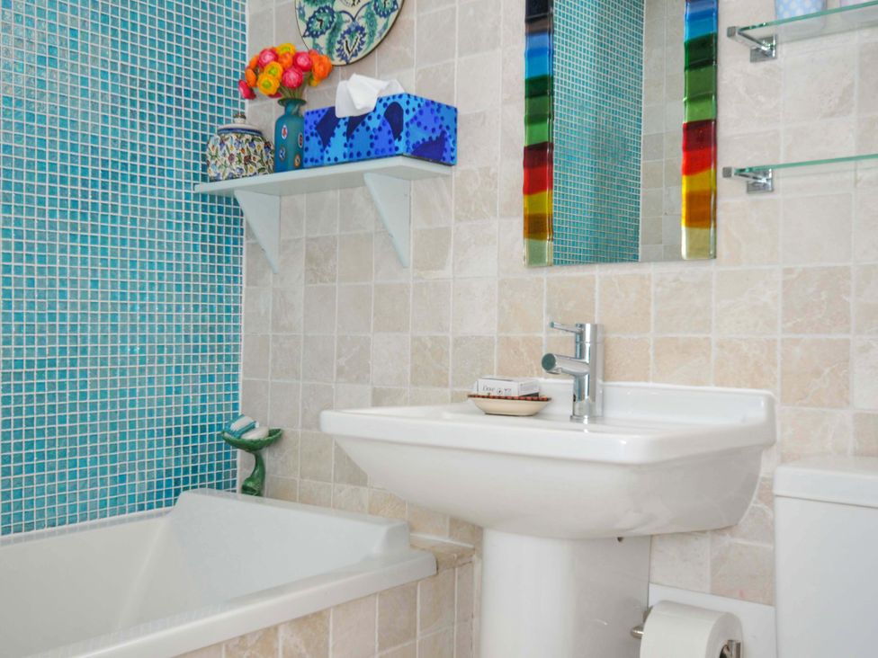 A bathroom with a white bathtub and sink blue tiled wall a colorful mirror and a shelf with flowers and a tissue box at 5 Esplanade in Fowey