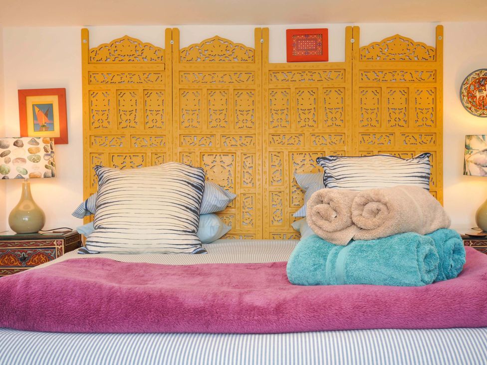 A bed with striped pillows and rolled towels in front of a wooden carved headboard with lamps and framed pictures on either side at 5 Esplanade in Fowey