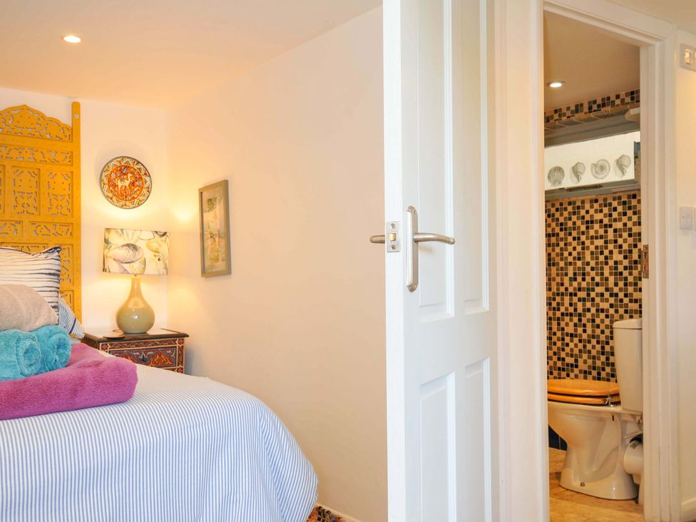 A bedroom with a bed with striped bedding and folded towels beside a bedside table with a lamp and decorative plate on the wall next to a partially open door leading to a bathroom with mosaic tiles and a toilet at 5 Esplanade in Fowey