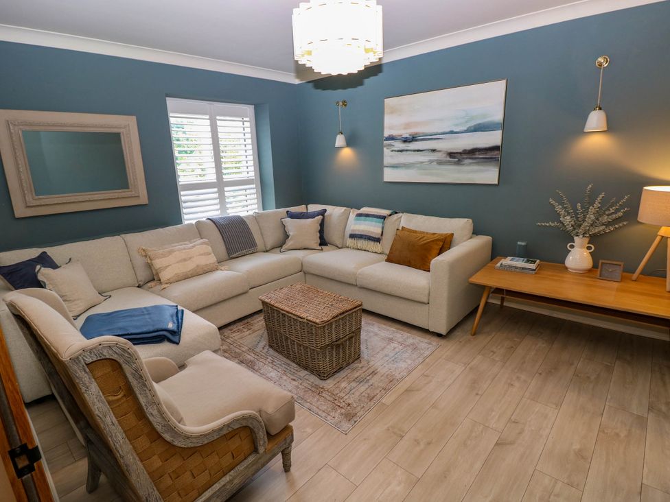 A living room with a large beige sectional sofa a wooden armchair a wicker basket coffee table a rug and a wooden console table with a lamp and vase at The Loft in Ystradgynlais