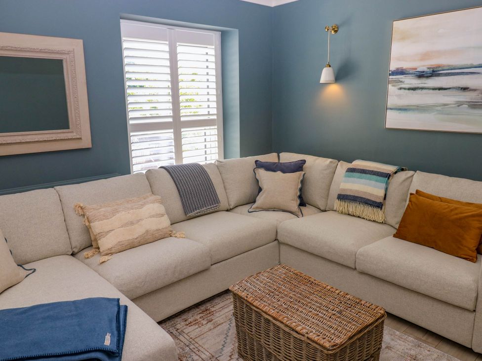 A living room with a beige sectional sofa with assorted pillows and blankets a wicker coffee table a window with white shutters a framed mirror on the wall and a wall light at The Loft in Ystradgynlais