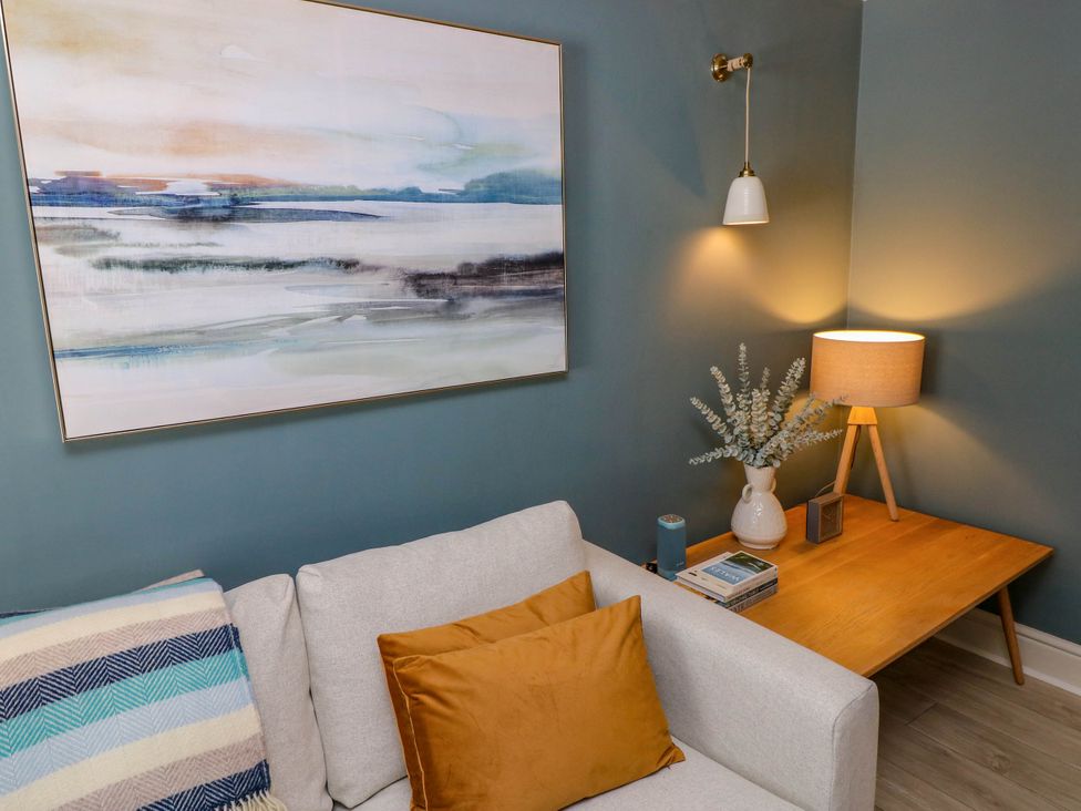 A living room corner with a light grey sofa with orange pillow and striped blanket next to a wooden side table with a lamp books clock and vase with plant at The Loft in Ystradgynlais
