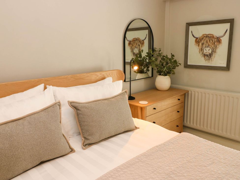A bedroom with a bed, pillows, a wooden bedside table with a plant, a lamp, a mirror, and a framed picture of a highland cow at The Loft in Ystradgynlais